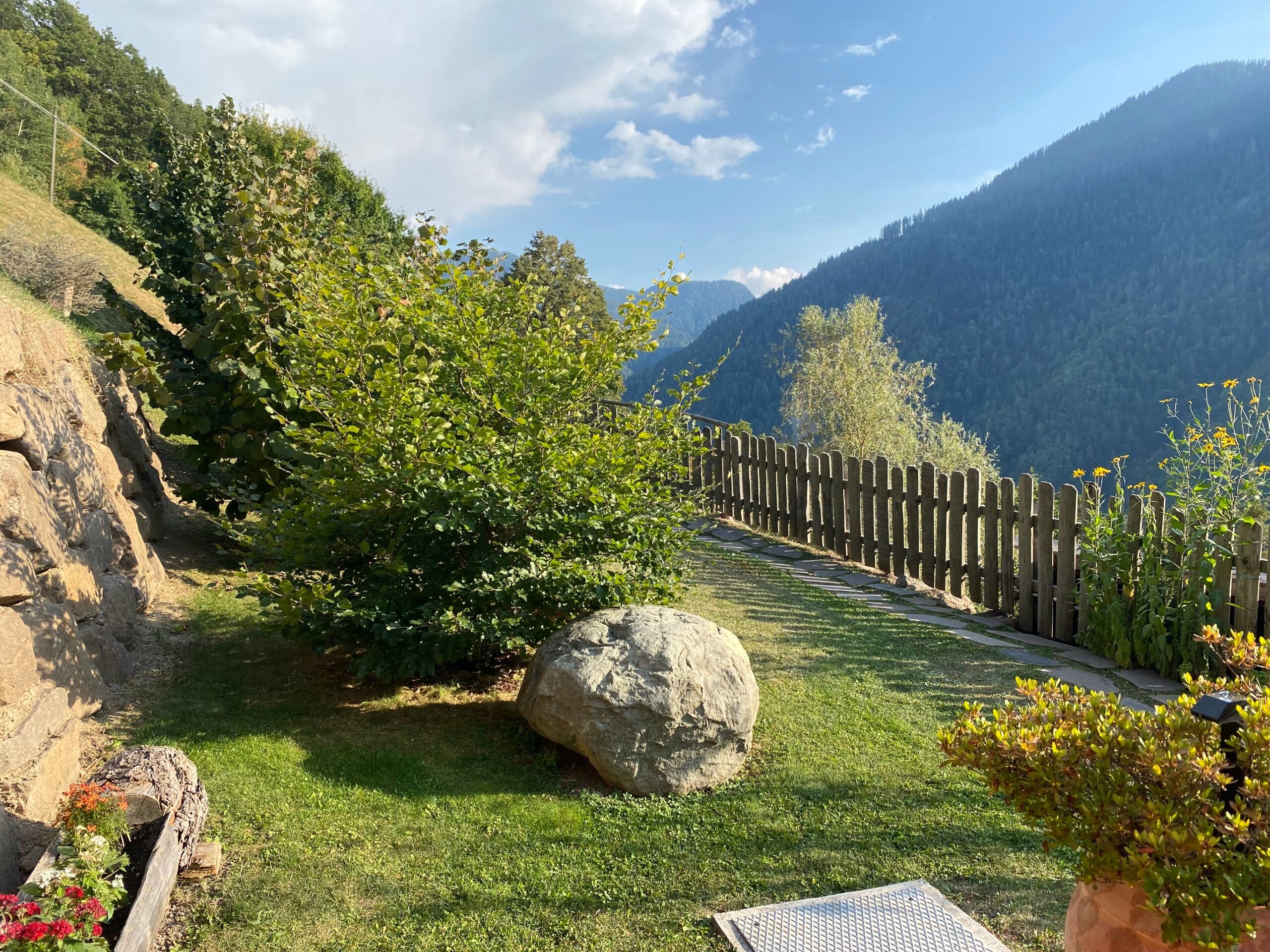 Garten mit großem Findling, Blumen und Holzzaun vor bewaldeten Bergen